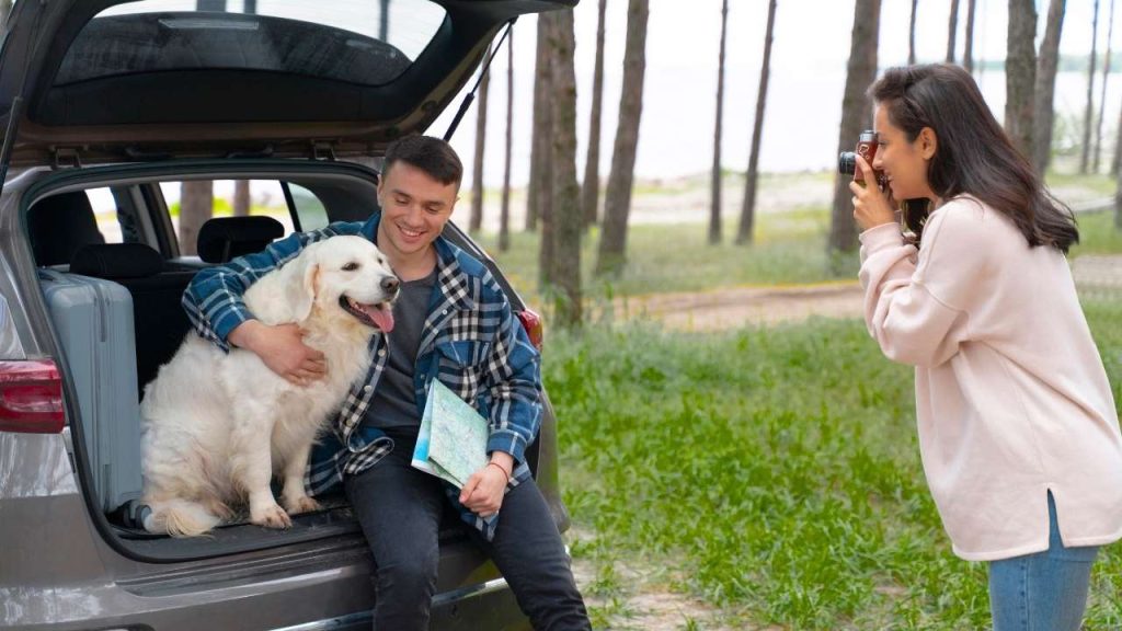 A woman takes a photo of a man and his happy golden retriever sitting in a car during a Montana road trip, representing pet insurance Montana.